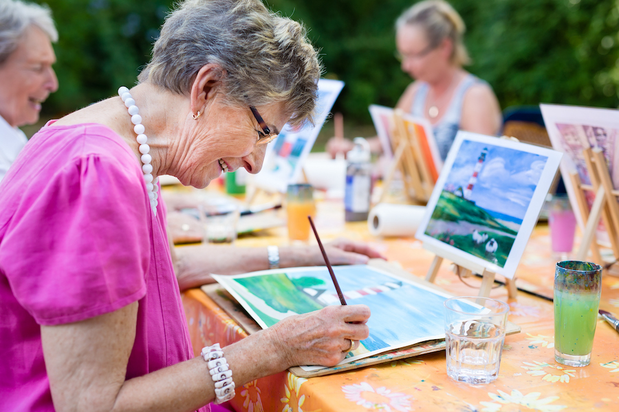 Side view of a happy senior woman smiling while painting with watercolors as a recreational activity outdoors at a retirement community with a group of other retired women